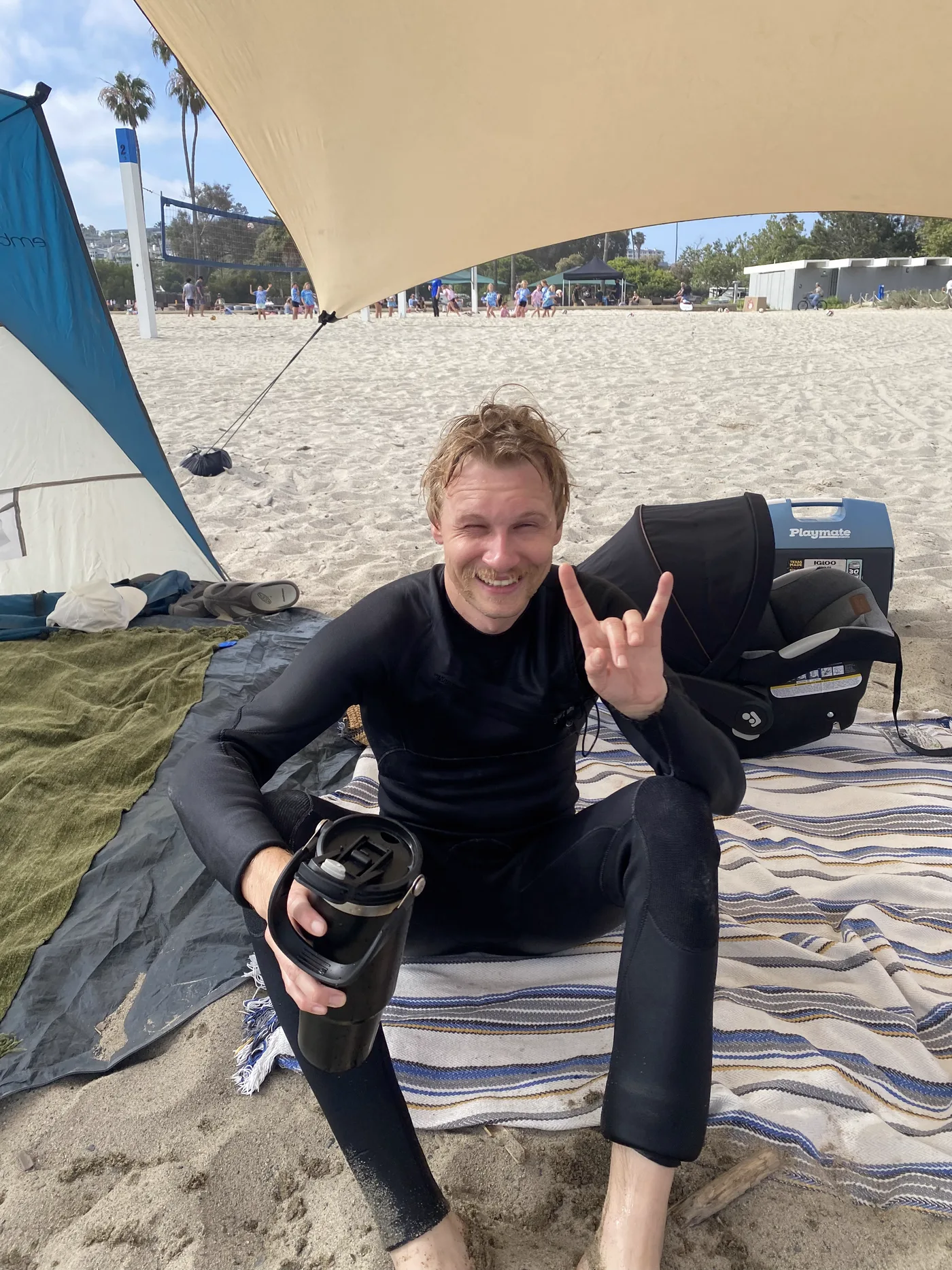 Cody at the beach in a wetsuit after surfing, grinning and throwing a hand sign